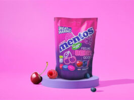 A pack of Mentos Feeling Berry Good sits on a purple coloured podium with pink walls around it. A raspberry also sits on a podium, with a cherry leaning on it and a berry rolling in front of it.