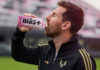 Lionel Messi drinks from a Mas+ by Messi Miami Punch drink in a football stadium.