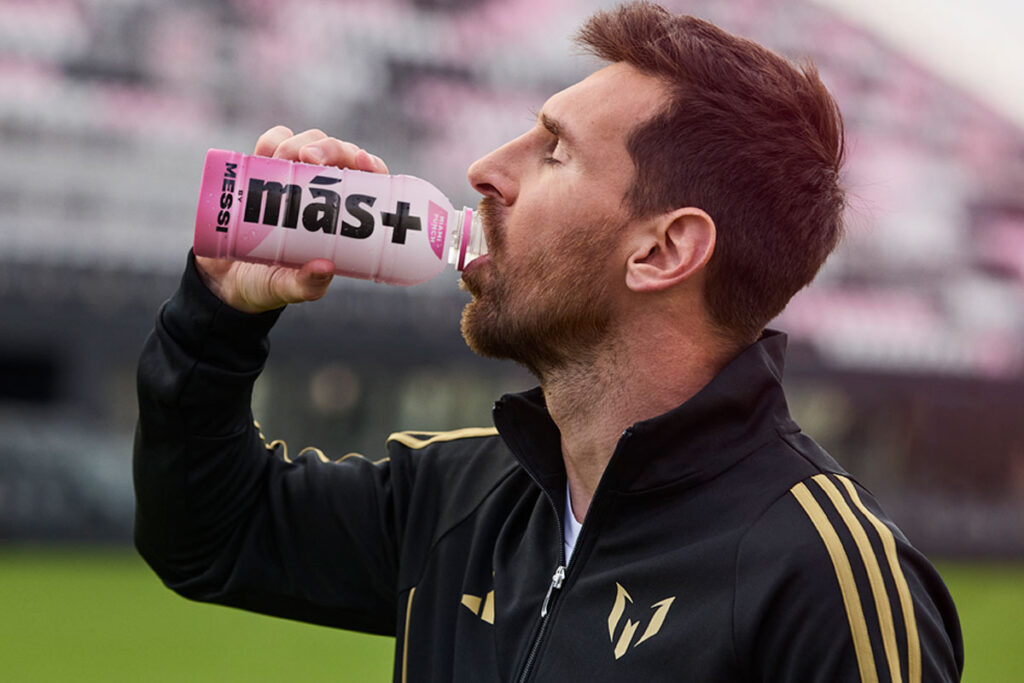 Lionel Messi drinks from a Mas+ by Messi Miami Punch drink in a football stadium.