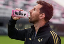 Lionel Messi drinks from a Mas+ by Messi Miami Punch drink in a football stadium.