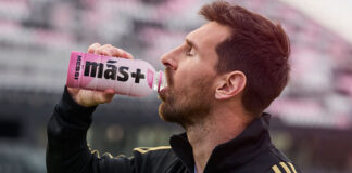 Lionel Messi drinks from a Mas+ by Messi Miami Punch drink in a football stadium.