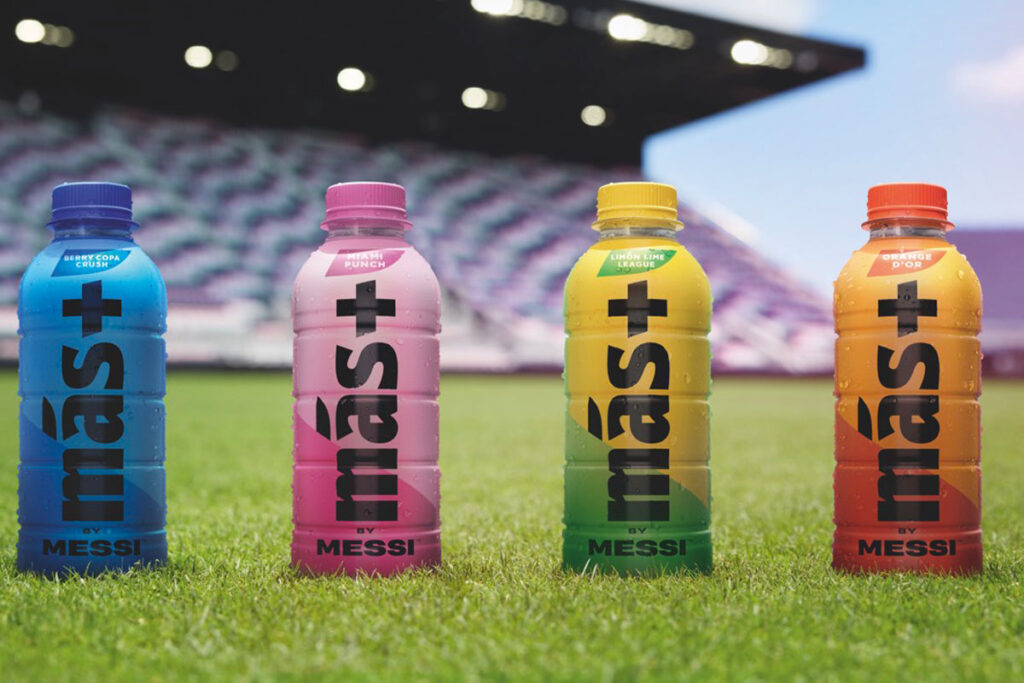 Bottles of Mas+ by Messi stand in the middle of a football ground with flavours lined up left to right: Berry Copa Crush, Miami Punch, Limon Lime League and Orange D'or.