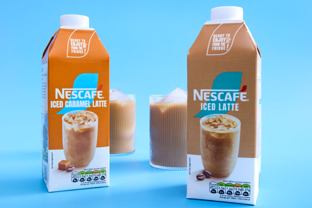 Cartons of Nescafé Iced Latte and Iced Caramel Latte stand against a blue background with the drinks poured out into ribbed glasses behind the cartons with ice.