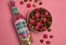 A bottle of Popcorn Kitchen Peter Pigs lies down on a pink background with a bowl that has chocolate covered popcorn and pieces of pig shaped strawberry chocolate with both the chocolate and popcorn lying around the bowl as well.