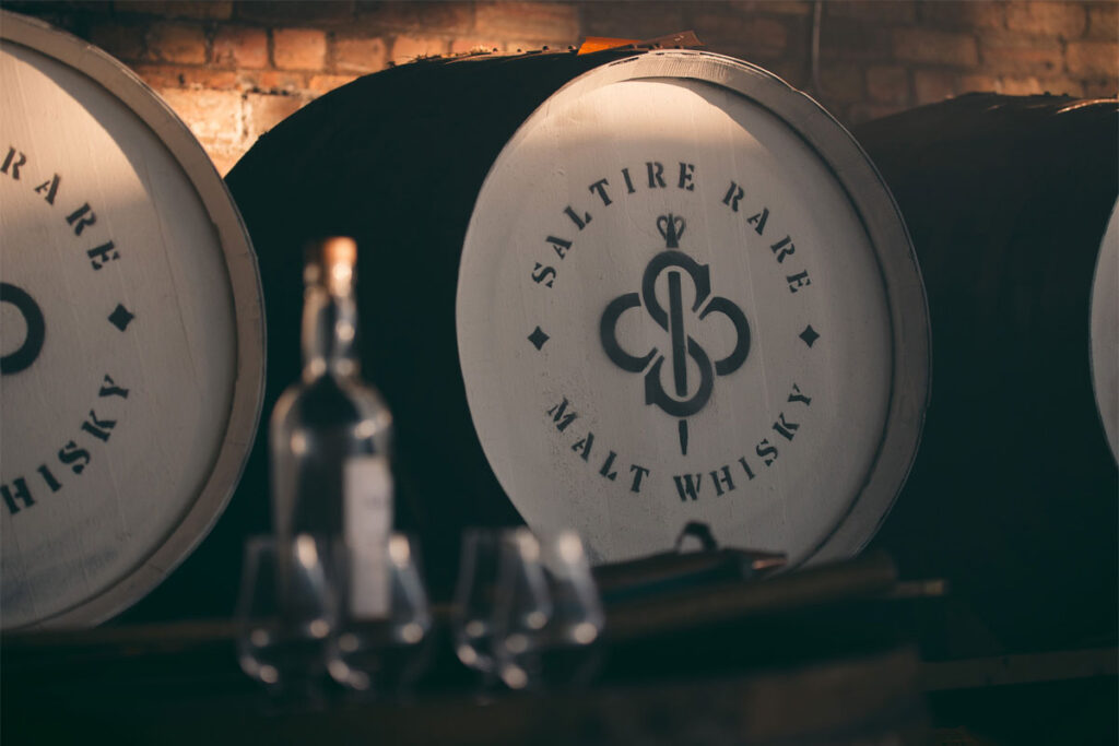 A whisky cask with the Saltire Rare Malt Whisky brand on the top of the lid with a bottle of whisky and nosing glasses in the foreground of the image.