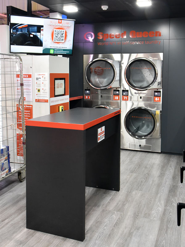 Washing machines in walls with a black counter with red lining in the middle of the floor with the Speed Queen logo on the wall above the washing machines.