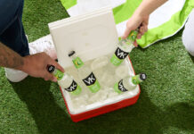 A red cooler filled with ice and bottles of VK Lemon & Lime are being picked out by people, the cooler is sitting on some grass with a green and white beach towel above it.