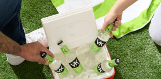 A red cooler filled with ice and bottles of VK Lemon & Lime are being picked out by people, the cooler is sitting on some grass with a green and white beach towel above it.