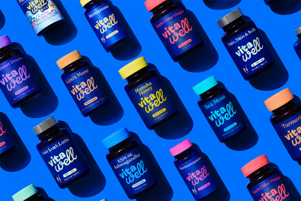 Bottles of Vitawell Vitamins and Supplements are lying down against a blue background, the bottles all have lids of varying colours to match the variant and the colour matches the text found on the bottle.