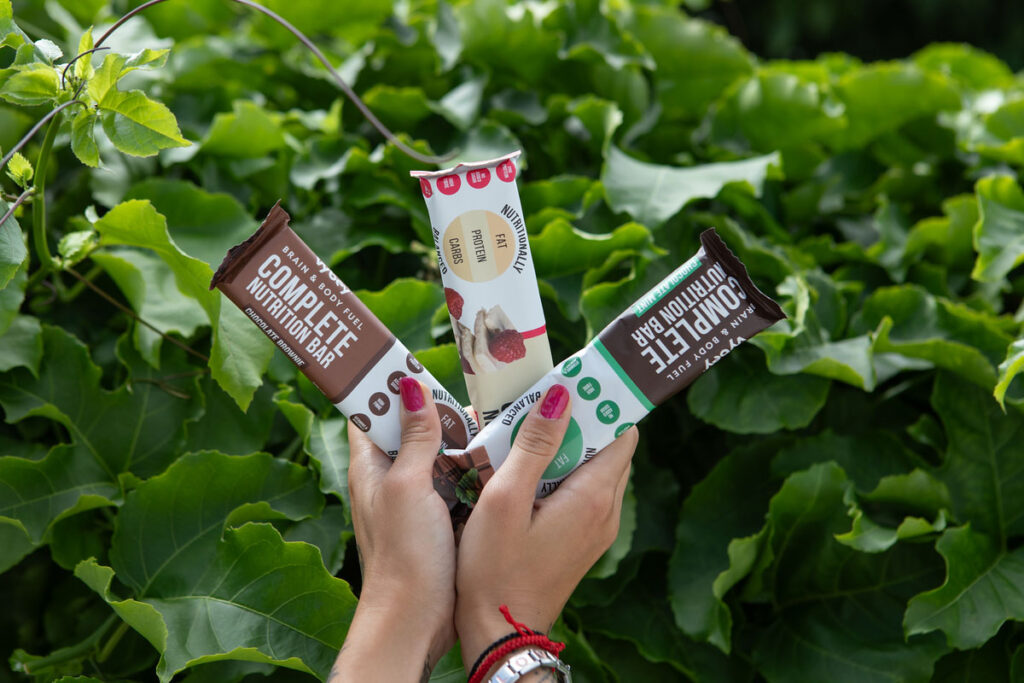 A person holds Vybey's three nutrition bars – Chocolate Brownie, Raspberry White Chocolate and Mint Chocolate – with a green bush behind the bars.