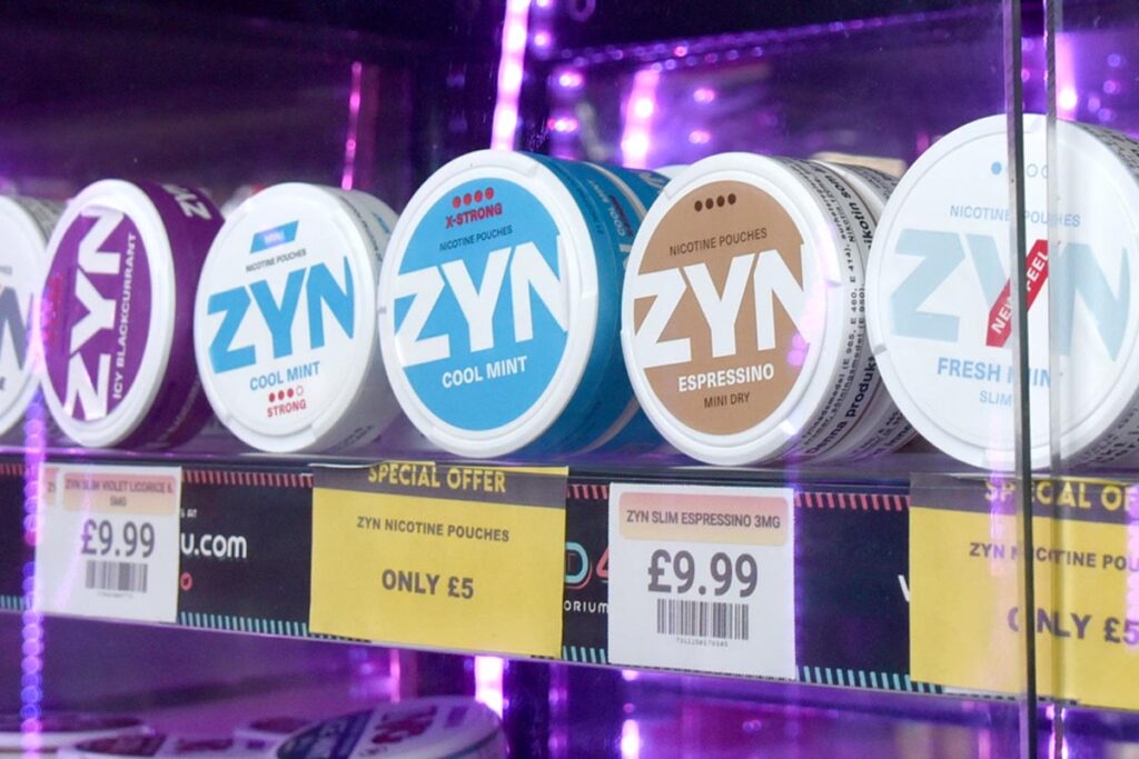 Packs of ZYN nicotine pouches are lined up in a glass cabinet, with yellow promotional price labels below them.