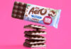 A bar of Aero Coconut Sharing is above halved pieces of the chocolate bar that have been stacked on top of each other against a pink background.