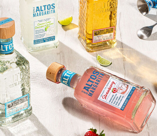 Altos launches Strawberry Margarita ready-to-serve variant A bottle of Altos Strawberry Margarita lies on its side on a wooden table with strawberries around the bottle. There is a bottle of Altos Tequila standing up to its left with a bottle of the Lime Margarita at the top of the image as well as a bottle of the Agave Tequila with lime segments, a barspoon and a measure.