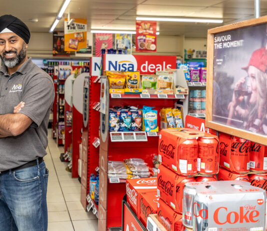 Coca-Cola celebrates convenience stores for 125th year in Great Britain A man stands with his arms folded to the left-hand side of the image of the inside of a convenience store with cans of Coca-Cola and Diet Coke stacked up on the right and promotional material above the cans.