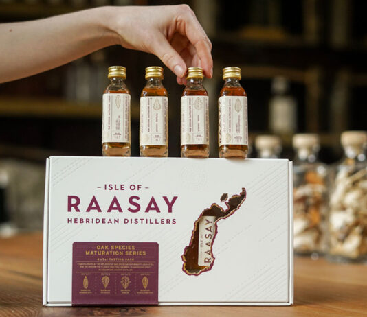 Explore whisky in new Isle of Raasay Tasting Pack The Isle of Raasay Oak Species Maturation Series Sampling Kit stands on a wooden table with sample bottles balanced on top of the kit as a hand reaches for the second bottle from the right. There are larger bottles blurred out in the background of the image.