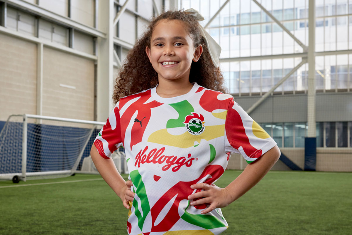 A young girl stands on a football pitch wearing the Kellogg's x Puma football top.