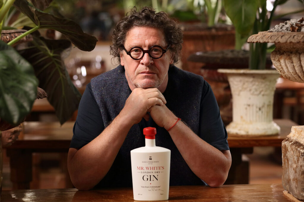 Marco Pierre White sits at a table with his hands resting under his chin with a bottle of Mr White's Gin on the table under his chin.