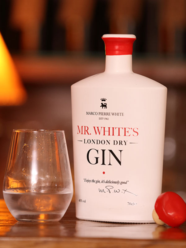 A bottle of Mr White's Gin is uncorked on a table with the cork sitting next to it and a glass with the gin inside.