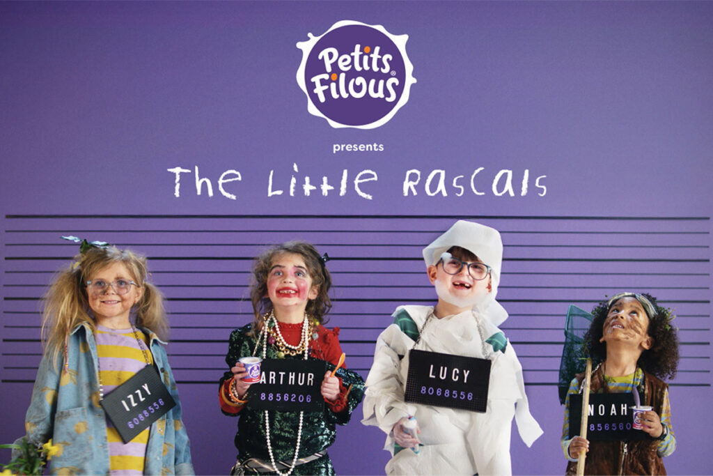 Four children are lined up against a police wall with purple walls as they are covered in mess as though they have been caught by their parents. Each holds a board with their name on it, on the wall behind them is the Petits Filous logo and the text 'The Little Rascals'.