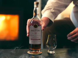Saltire introduces its oldest whisky bottling A bottle of Saltire Single Malt Whisky 18 Years Old is on a table as a person goes to grab the bottle, there is a nosing glass to the right of the bottle and a fireplace in the background to the left of the bottle.
