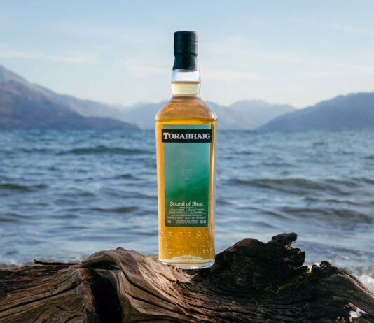 Torabhaig Skye whisky unveils fourth expression A bottle of Torabhaig Sound of Sleet is balanced on a wooden log with the sea in the background and hills on the landscape.