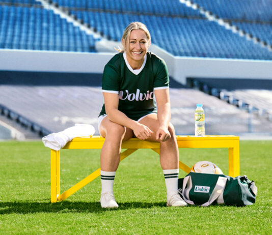 Volvic announced as Official Water Supplier of Women’s Rugby World Cup Natasha Hunt, English Rugby Union player and brand ambassador, sits on a yellow bench in the middle of a rugby ground with a bottle of Volvic on the bench to her right, a towel to her left and a kit bag next to her left foot.