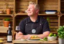 Matthew 'Hoggy' Hoggard stands in a garden area in front of a cooking table with food and a chopping board on top of it. There is a a bottle of Andrew Peace Shiraz on the table that has also been poured into a wine glass next to the bottle.