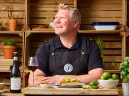 Matthew 'Hoggy' Hoggard stands in a garden area in front of a cooking table with food and a chopping board on top of it. There is a a bottle of Andrew Peace Shiraz on the table that has also been poured into a wine glass next to the bottle.