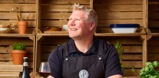 Matthew 'Hoggy' Hoggard stands in a garden area in front of a cooking table with food and a chopping board on top of it. There is a a bottle of Andrew Peace Shiraz on the table that has also been poured into a wine glass next to the bottle.