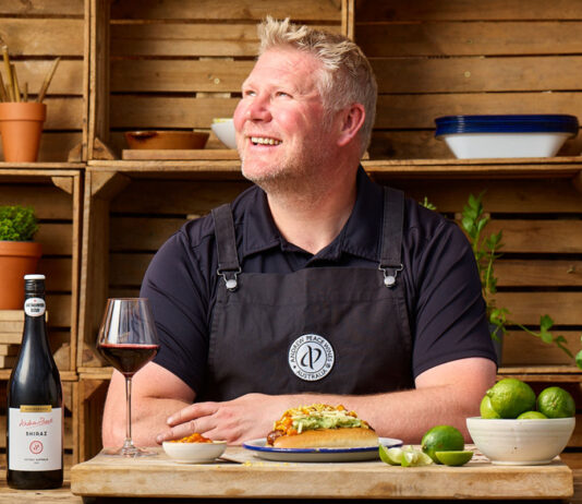 Andrew Peace wine partners with cricket legend Matthew Hoggard Matthew 'Hoggy' Hoggard stands in a garden area in front of a cooking table with food and a chopping board on top of it. There is a a bottle of Andrew Peace Shiraz on the table that has also been poured into a wine glass next to the bottle.