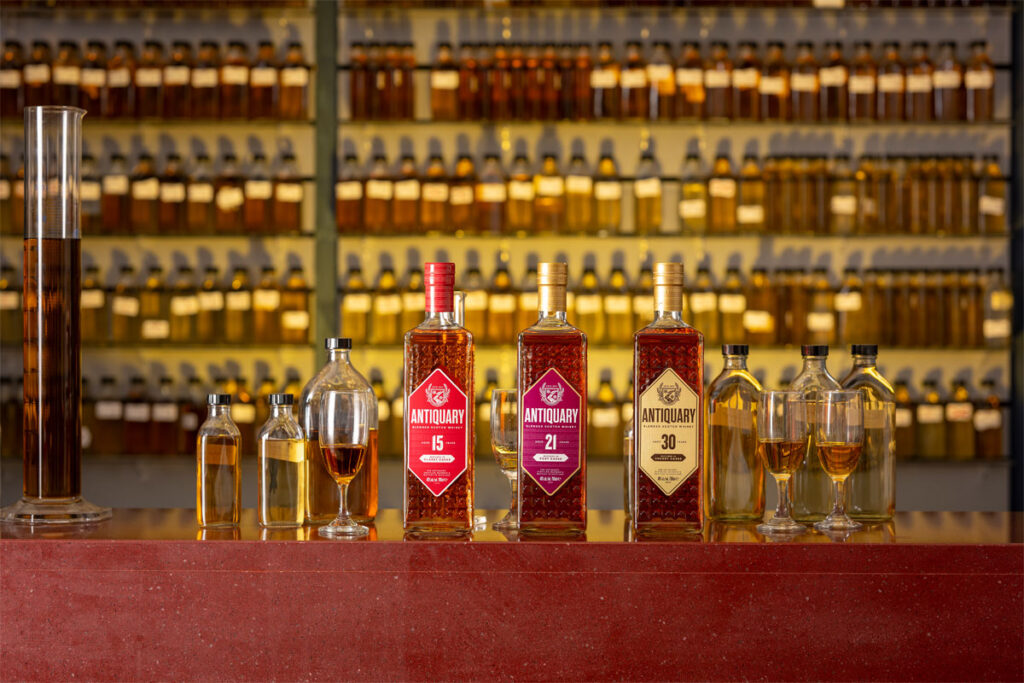 Bottles of The Antiquary whisky are lined up on a bar featuring the 15 Year Old, 21 Year Old and 30 Year Old. Along with these bottles, there are smaller sample bottles for blending as well as whisky glasses to serve in with unlabelled whisky sample bottles on shelves behind the bar.