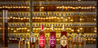 Bottles of The Antiquary whisky are lined up on a bar featuring the 15 Year Old, 21 Year Old and 30 Year Old. Along with these bottles, there are smaller sample bottles for blending as well as whisky glasses to serve in with unlabelled whisky sample bottles on shelves behind the bar.