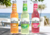 Bottles of Breezer consisting Zesty Orange, Zingy Lime and Crisp Watermelon all stand on a wooden table with a beach scene in the background behind the bottles.