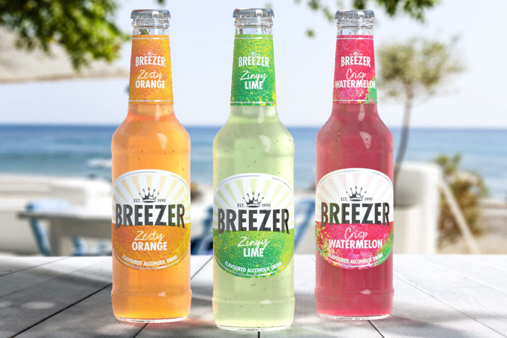 Bottles of Breezer consisting Zesty Orange, Zingy Lime and Crisp Watermelon all stand on a wooden table with a beach scene in the background behind the bottles.