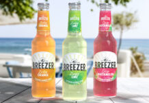 Bottles of Breezer consisting Zesty Orange, Zingy Lime and Crisp Watermelon all stand on a wooden table with a beach scene in the background behind the bottles.