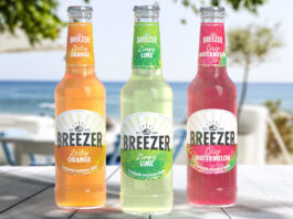 Bacardi Breezer makes its official comeback Bottles of Breezer consisting Zesty Orange, Zingy Lime and Crisp Watermelon all stand on a wooden table with a beach scene in the background behind the bottles.