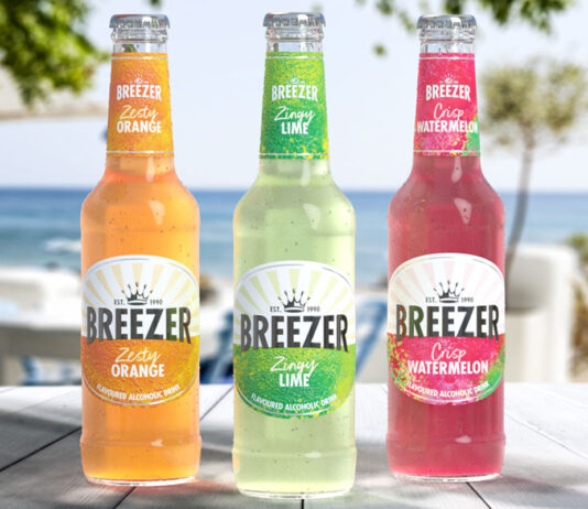 Bacardi Breezer makes its official comeback Bottles of Breezer consisting Zesty Orange, Zingy Lime and Crisp Watermelon all stand on a wooden table with a beach scene in the background behind the bottles.