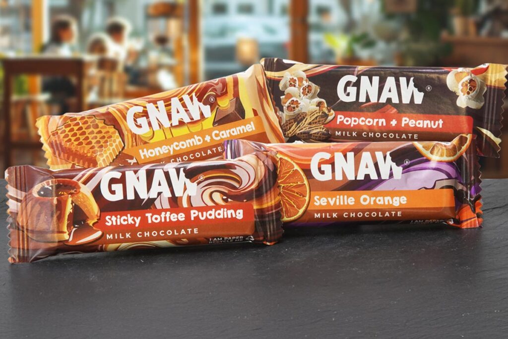 Packs of Gnaw impulse chocolate bars are stacked on a countertop with a cafe scene in the background. Variants include Honeycomb + Caramel, Popcorn + Peanut, Sticky Toffee Pudding and Seville Orange.
