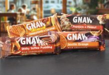 Packs of Gnaw impulse chocolate bars are stacked on a countertop with a cafe scene in the background. Variants include Honeycomb + Caramel, Popcorn + Peanut, Sticky Toffee Pudding and Seville Orange.