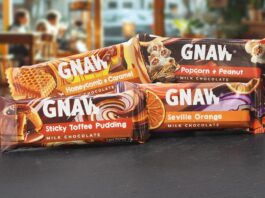 Packs of Gnaw impulse chocolate bars are stacked on a countertop with a cafe scene in the background. Variants include Honeycomb + Caramel, Popcorn + Peanut, Sticky Toffee Pudding and Seville Orange.