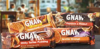 Packs of Gnaw impulse chocolate bars are stacked on a countertop with a cafe scene in the background. Variants include Honeycomb + Caramel, Popcorn + Peanut, Sticky Toffee Pudding and Seville Orange.