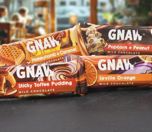 GNAW chocolate brings a four-strong impulse range to the table Packs of Gnaw impulse chocolate bars are stacked on a countertop with a cafe scene in the background. Variants include Honeycomb + Caramel, Popcorn + Peanut, Sticky Toffee Pudding and Seville Orange.