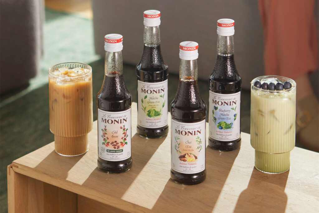 Bottles of Monin Concentrate variants including Cold Brew, Matcha Green Tea, Chai Tea and Blueberry Matcha Green Tea with iced coffee serves in the glasses around them.