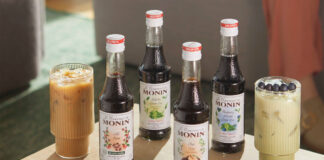 Bottles of Monin Concentrate variants including Cold Brew, Matcha Green Tea, Chai Tea and Blueberry Matcha Green Tea with iced coffee serves in the glasses around them.