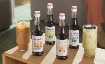 Bottles of Monin Concentrate variants including Cold Brew, Matcha Green Tea, Chai Tea and Blueberry Matcha Green Tea with iced coffee serves in the glasses around them.