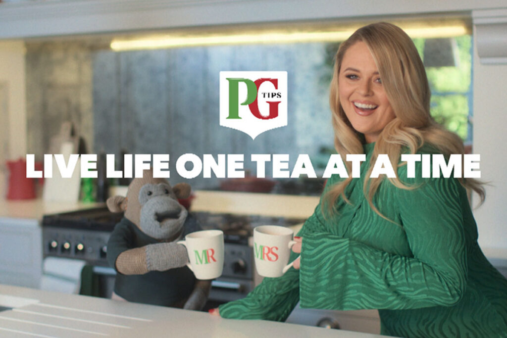The PG Tips Monkey mascot with comic actor Emily Atack stand in a kitchen with Mr and Mrs Mugs as they go to clink them together. The PG Tips logo is on top of the image with text that reads 'LIVE LIFE ONE TEA AT A TIME'.