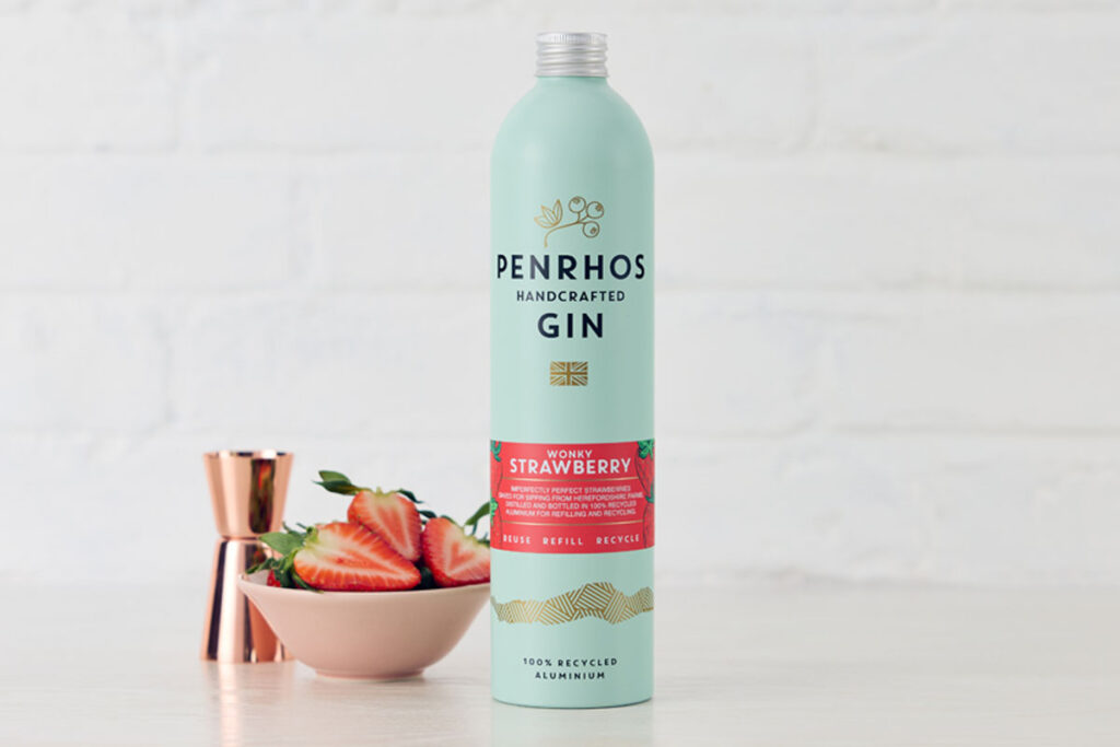 A bottle of Penrhos Handcrafted Wonky Strawberry Gin stands on a white table against a white background with a pink bowl of strawberries next to the bottle and a rose gold jigger measure behind the bowl.
