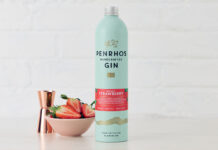 A bottle of Penrhos Handcrafted Wonky Strawberry Gin stands on a white table against a white background with a pink bowl of strawberries next to the bottle and a rose gold jigger measure behind the bowl.