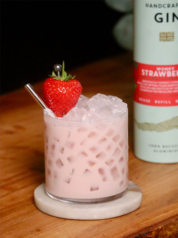 A Penrhos Strawberries & Cream Cocktail is served in a rocks glass with a bottle of Penrhos Wonky Strawberry Gin behind it.