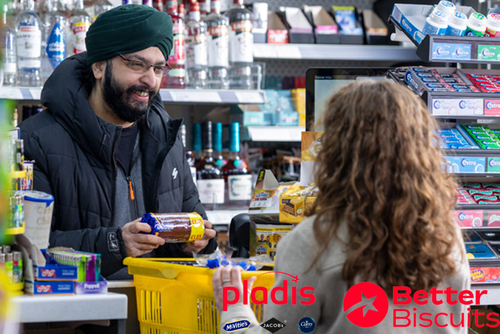 A man serves a customer at a till point inside a convenience store. The customer has placed a yellow shopping basket onto the counter with items inside it as the retailer takes items out to scan them. The retailer is holding a packet of McVitie's Chocolate Digestives.